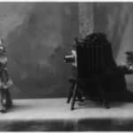 A cat is posed seated on a chair in front of another cat operating a camera.