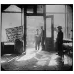 Three men stand at the door of an empty store front. A wagon out front is loaded with large sacks. A sign in the window in English and Yiddish reads 'Help the garment workers in their fight for bread and freedom.'