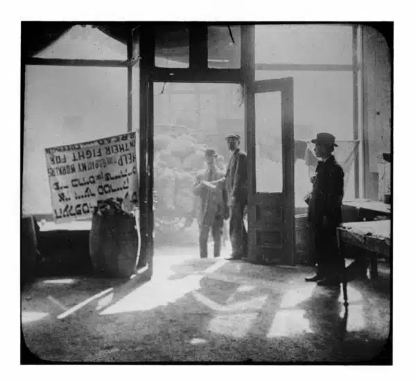 Three men stand at the door of an empty store front. A wagon out front is loaded with large sacks. A sign in the window in English and Yiddish reads 'Help the garment workers in their fight for bread and freedom.'